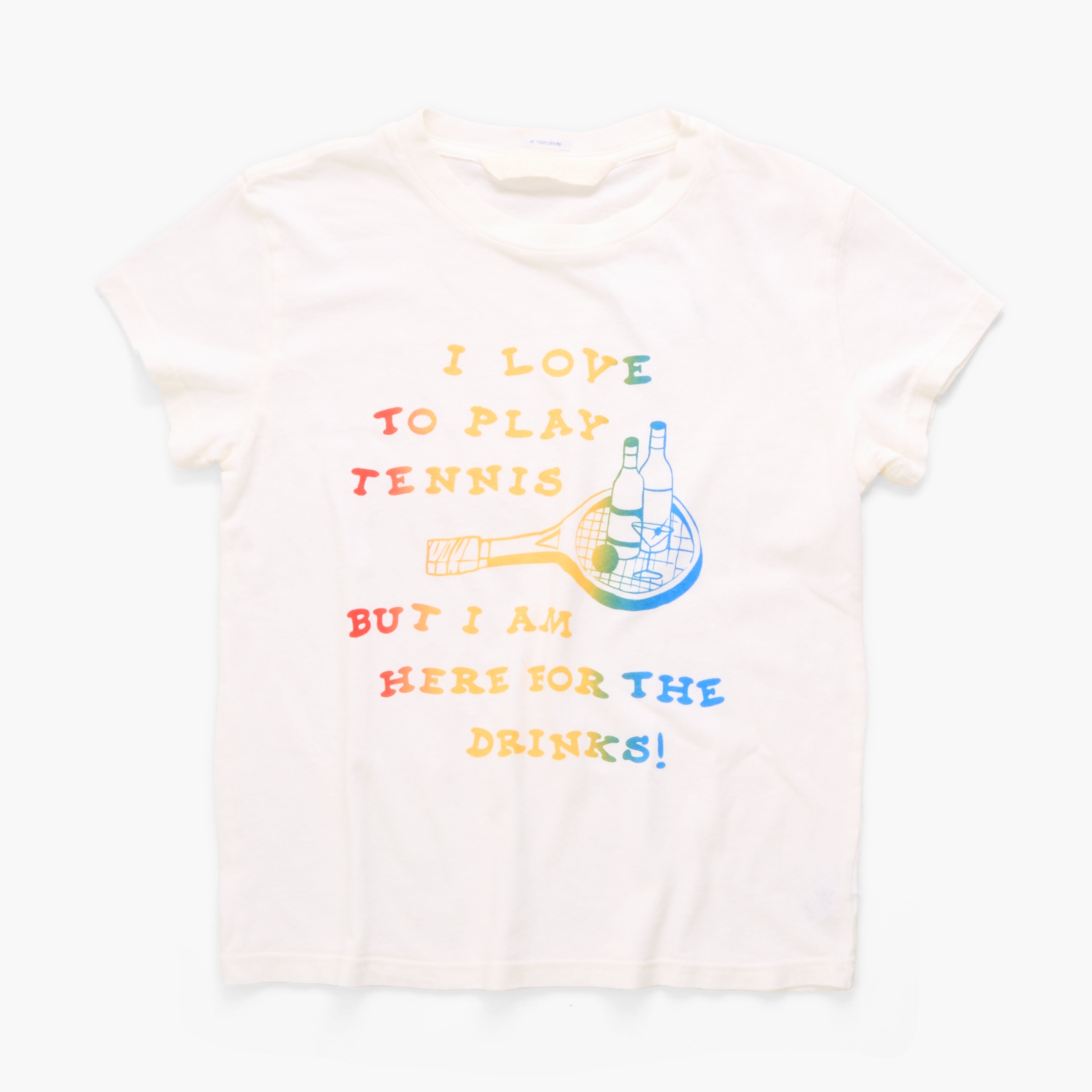 A white short-sleeve t-shirt with a colorful graphic print on the front. The graphic features the text 'I LOVE TO PLAY TENNIS BUT I AM HERE FOR THE DRINKS!' in a rainbow gradient, alongside an illustration of a tennis racket and two beer bottles.