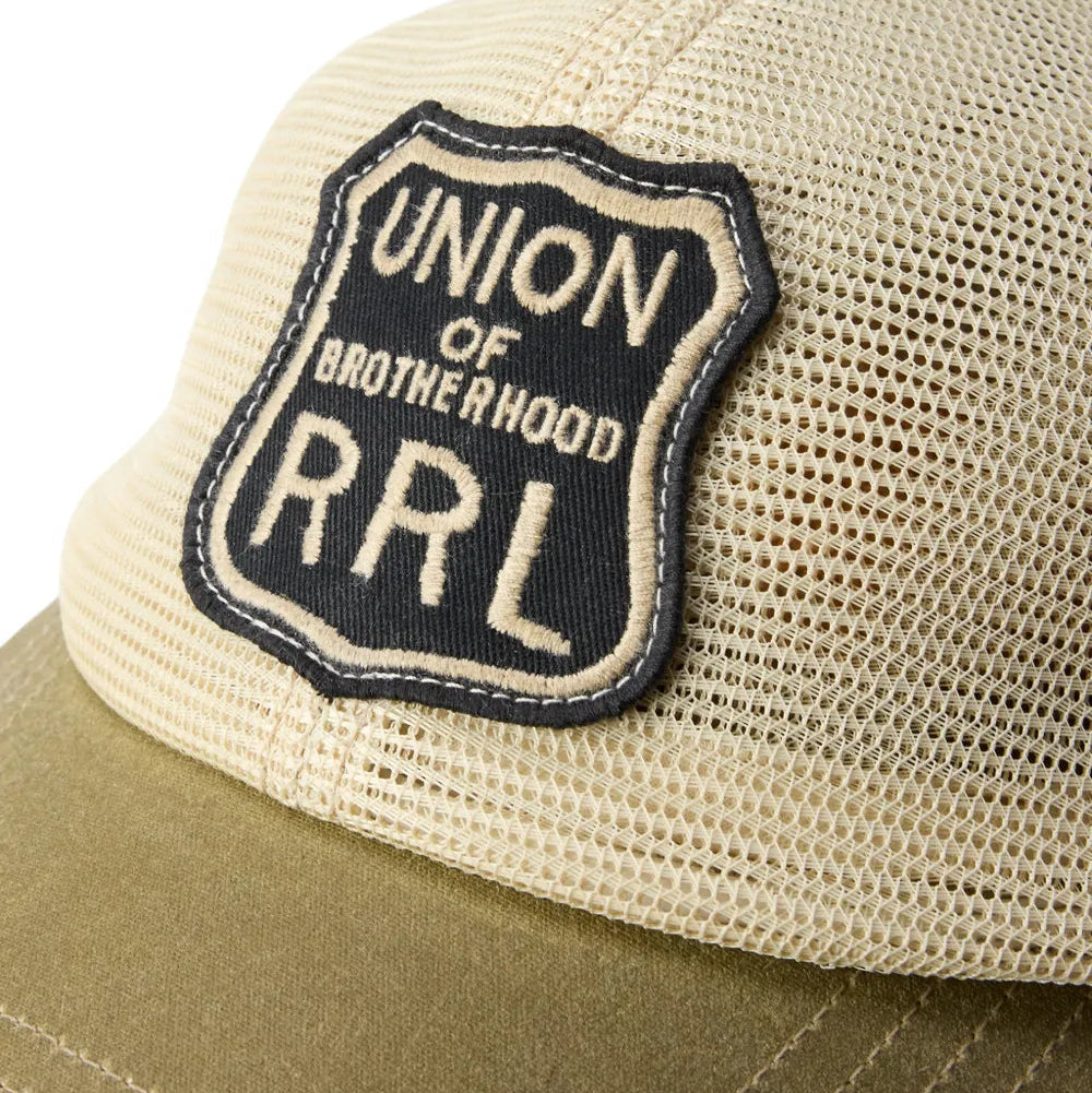 Close-up of a trucker cap with a beige mesh crown, olive green brim, and a black shield-shaped patch reading 'UNION OF BROTHERHOOD RRL'.