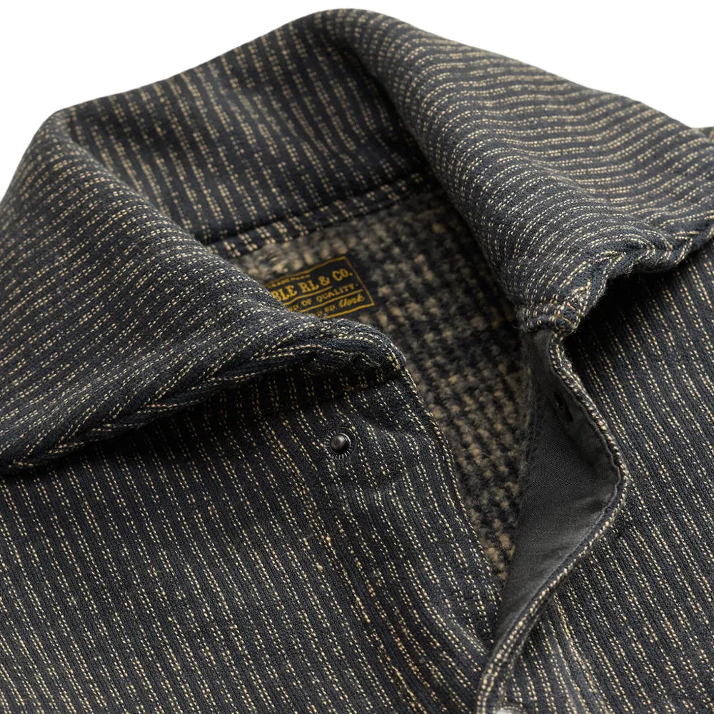 Close-up of a dark charcoal gray and black striped textured overshirt with a pointed collar and a visible yellow brand label inside the collar.