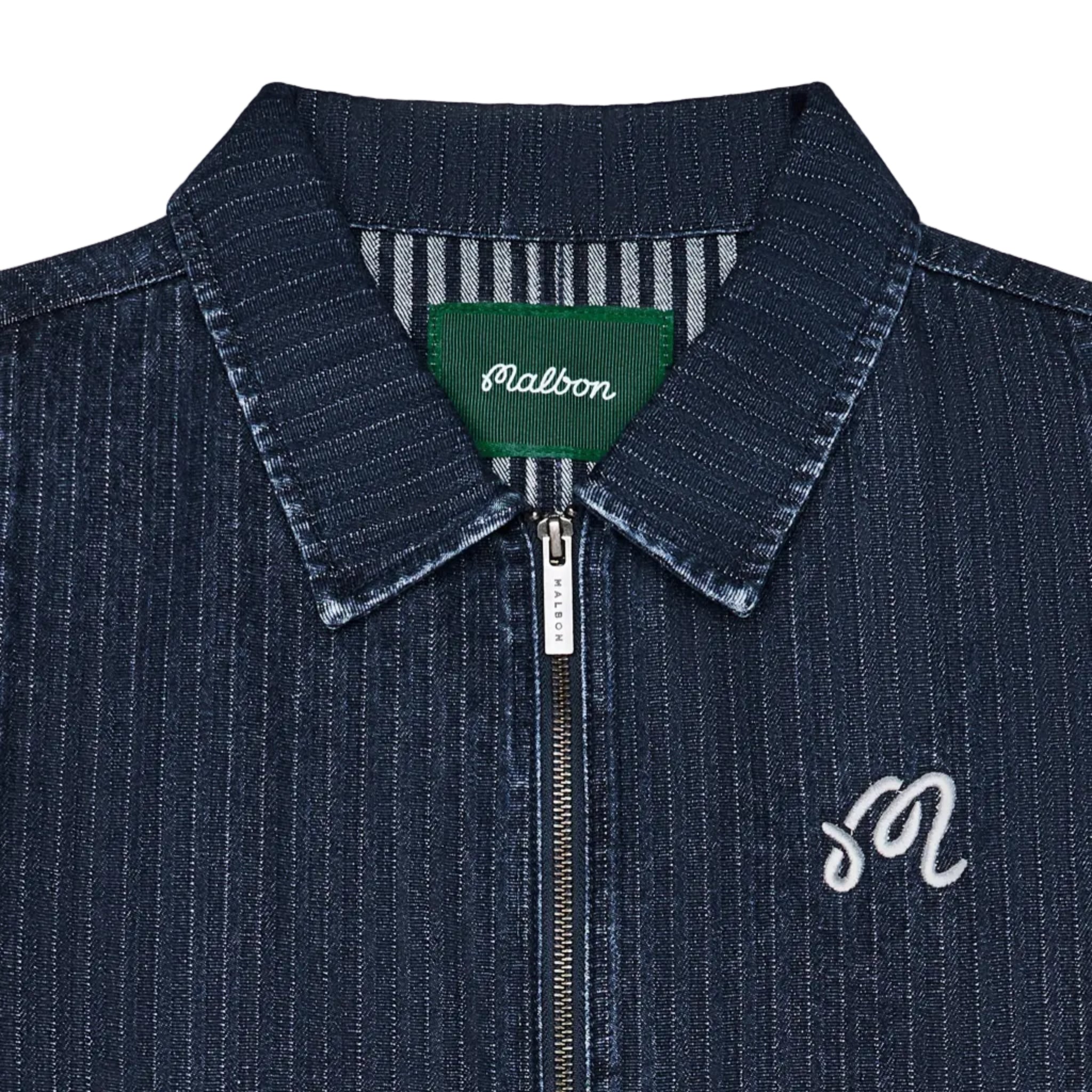 Close-up of a dark blue denim zip-up jacket with a pointed collar, featuring a green Malbon neck label and a white embroidered script 'M' on the chest.
