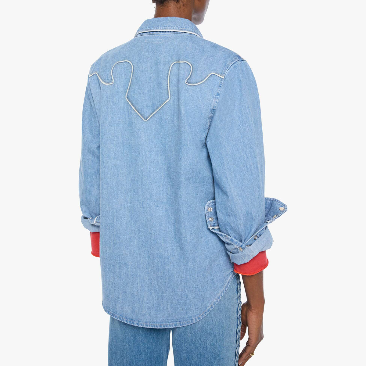 A person shown from the back wearing a light blue denim shirt jacket with white contrast stitching and rolled sleeves, paired with blue jeans against a plain light background.