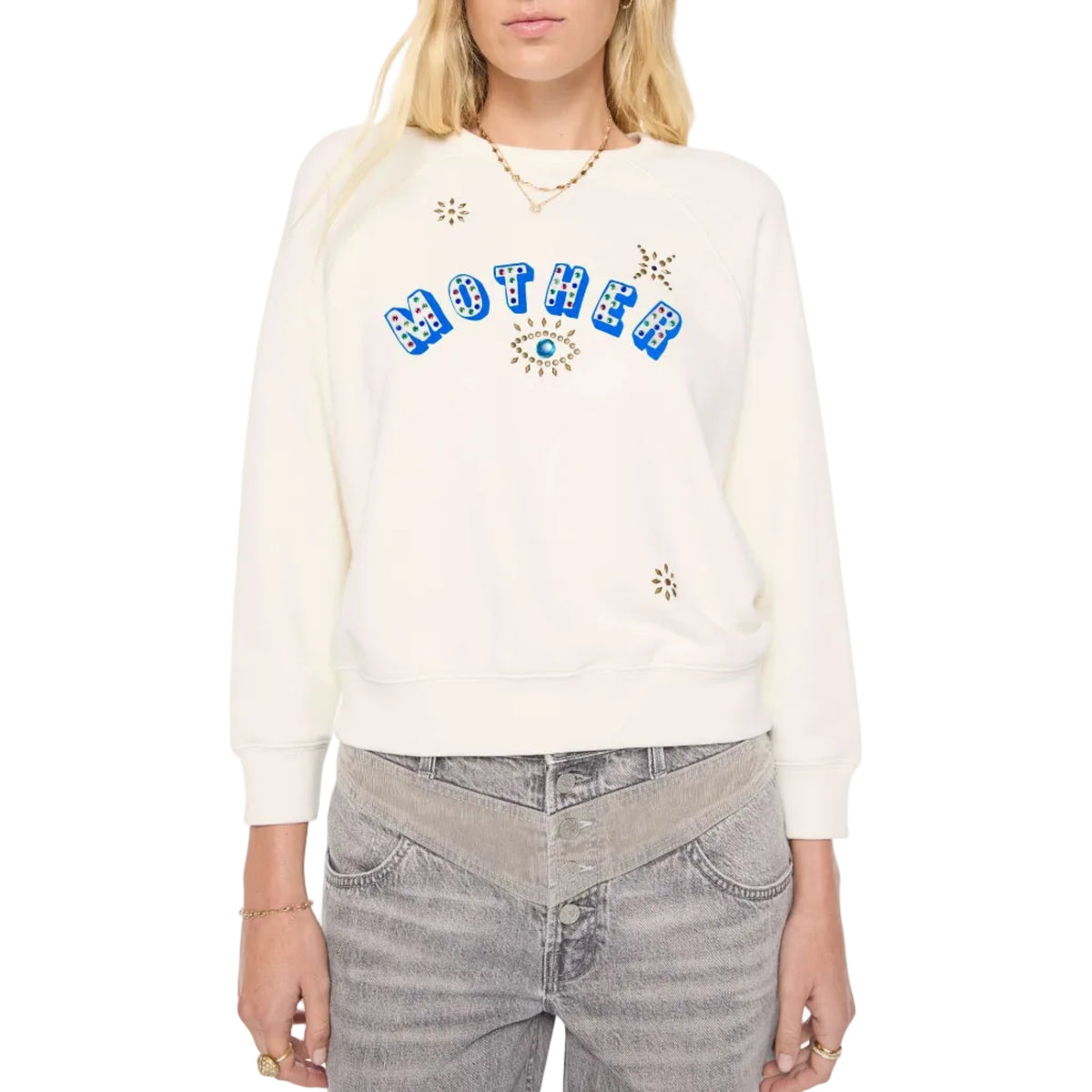 A woman modeling a cream-colored sweatshirt with 'MOTHER' written in blue letters across the chest, embellished with small stars and an eye motif against a plain white background.