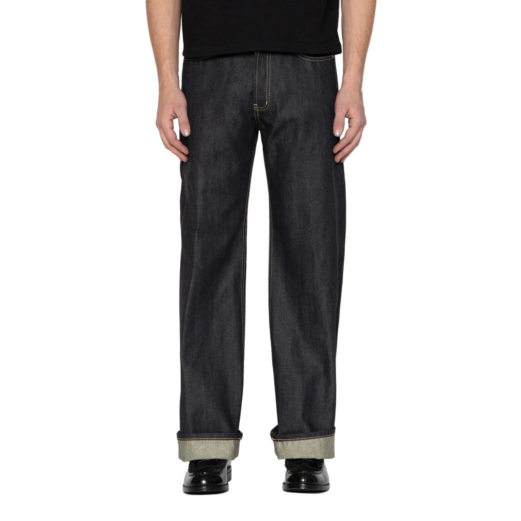A person from the waist down wearing dark indigo straight-leg jeans with a cuffed hem, paired with a black t-shirt and black shoes, against a white background.