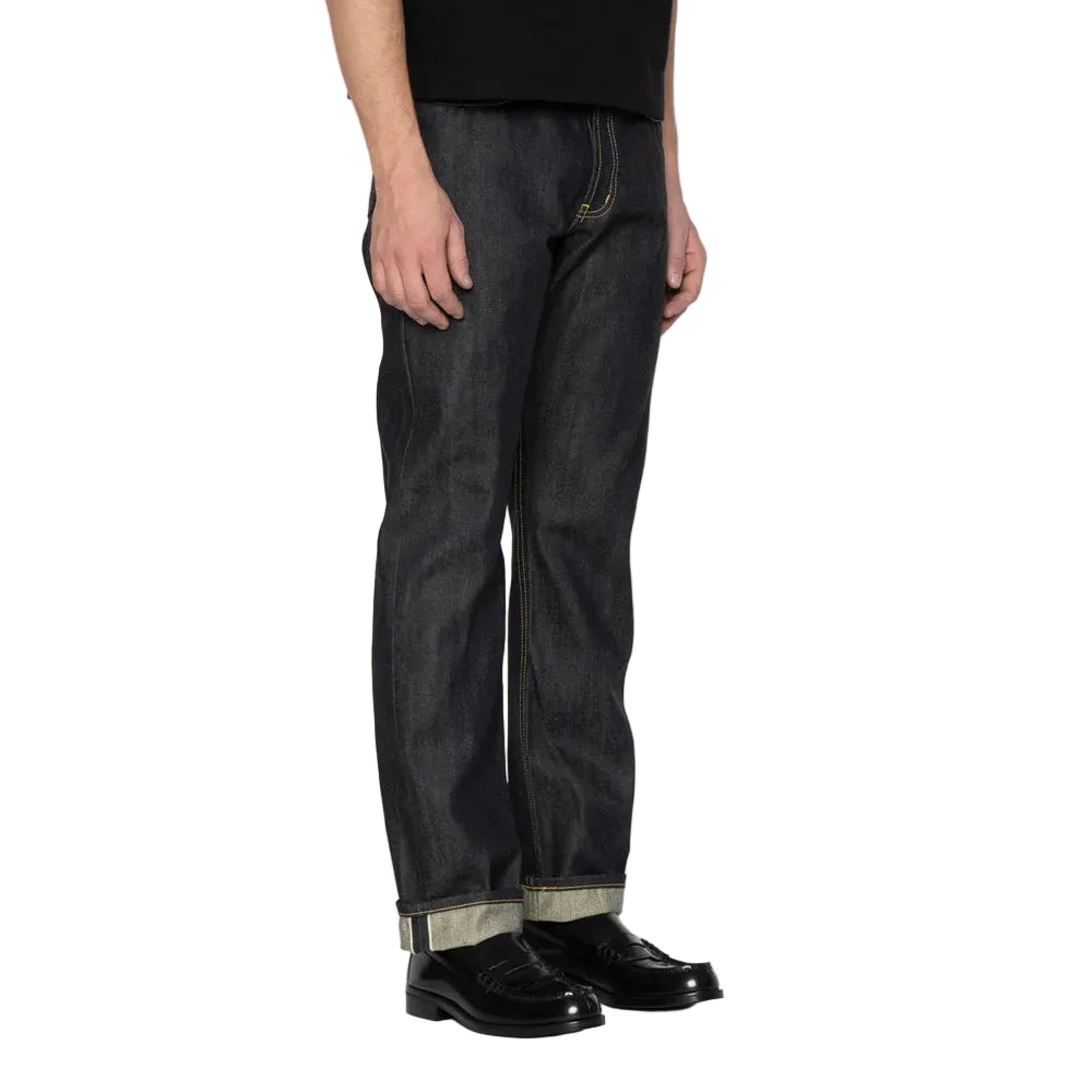 A person wearing dark indigo denim jeans with rolled cuffs showing a light inner lining, paired with black leather shoes, photographed against a white background.