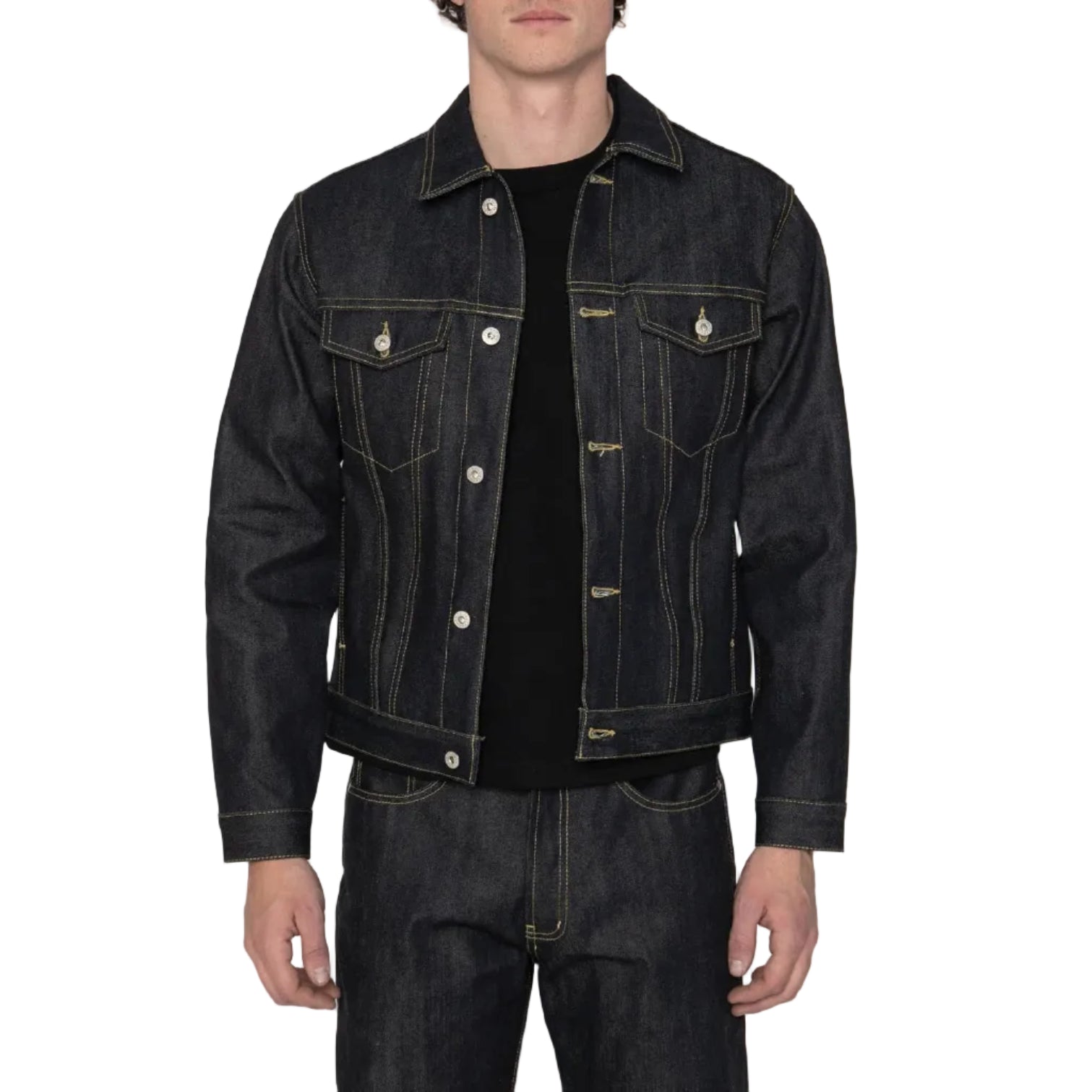 A man wearing a dark indigo denim trucker jacket over a black t-shirt and matching dark jeans, standing against a white background.