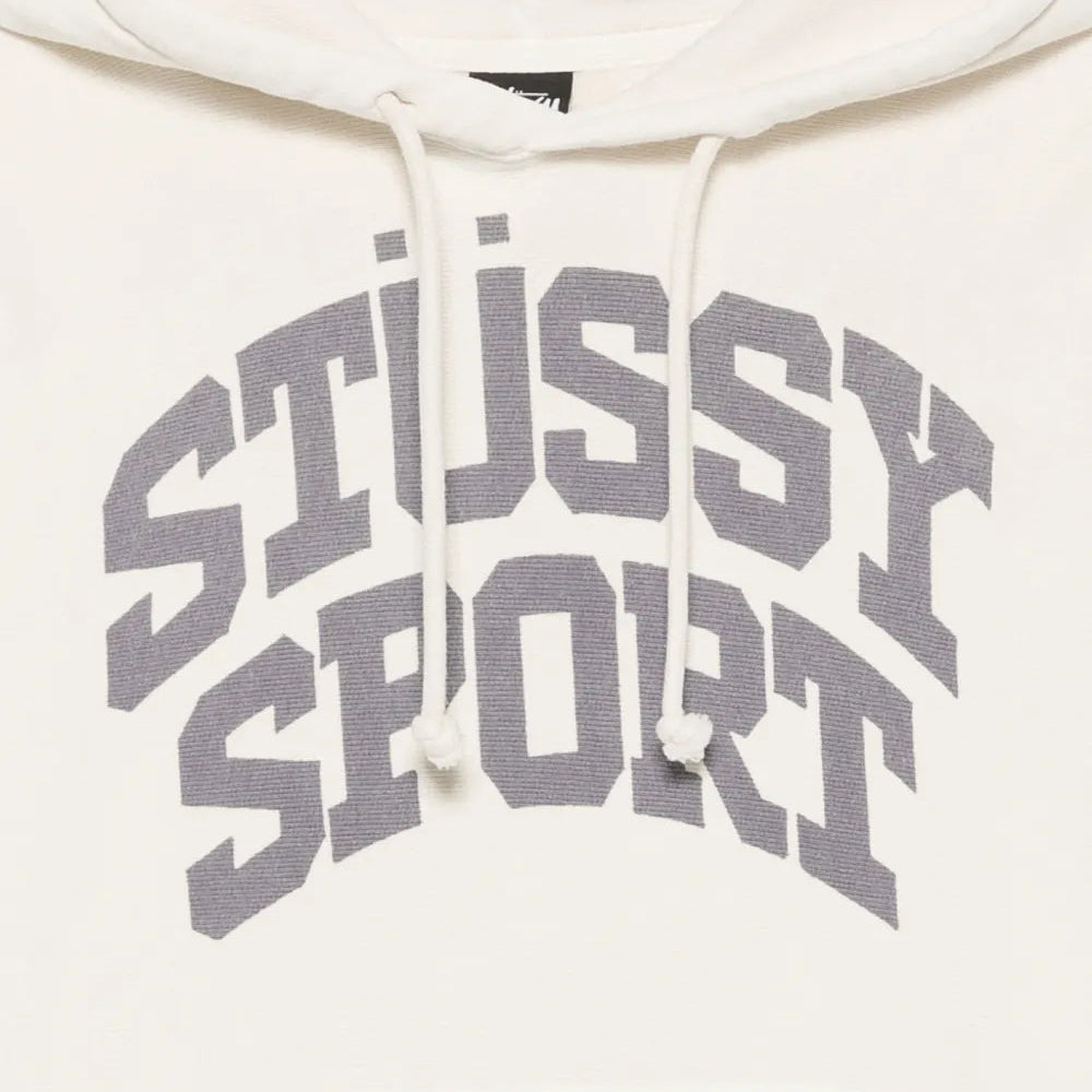 Close-up of a cream-colored hoodie featuring a large gray arched 'STUSSY SPORT' logo printed across the chest, with white drawstrings.