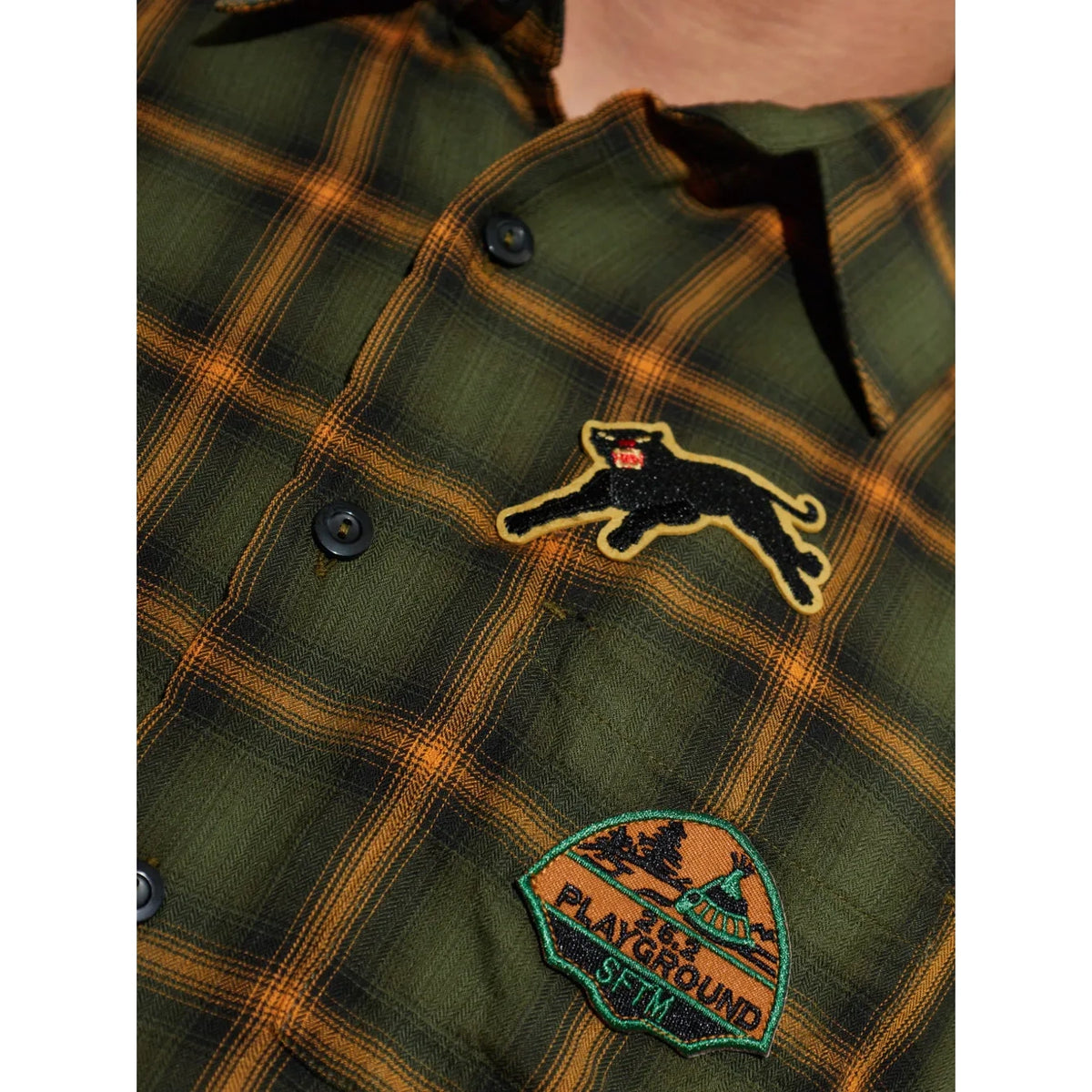 Close-up of the School Shirt’s chest, showing the embroidered black panther patch and the “26.2 Playground SFTM” crest patch on the plaid fabric.