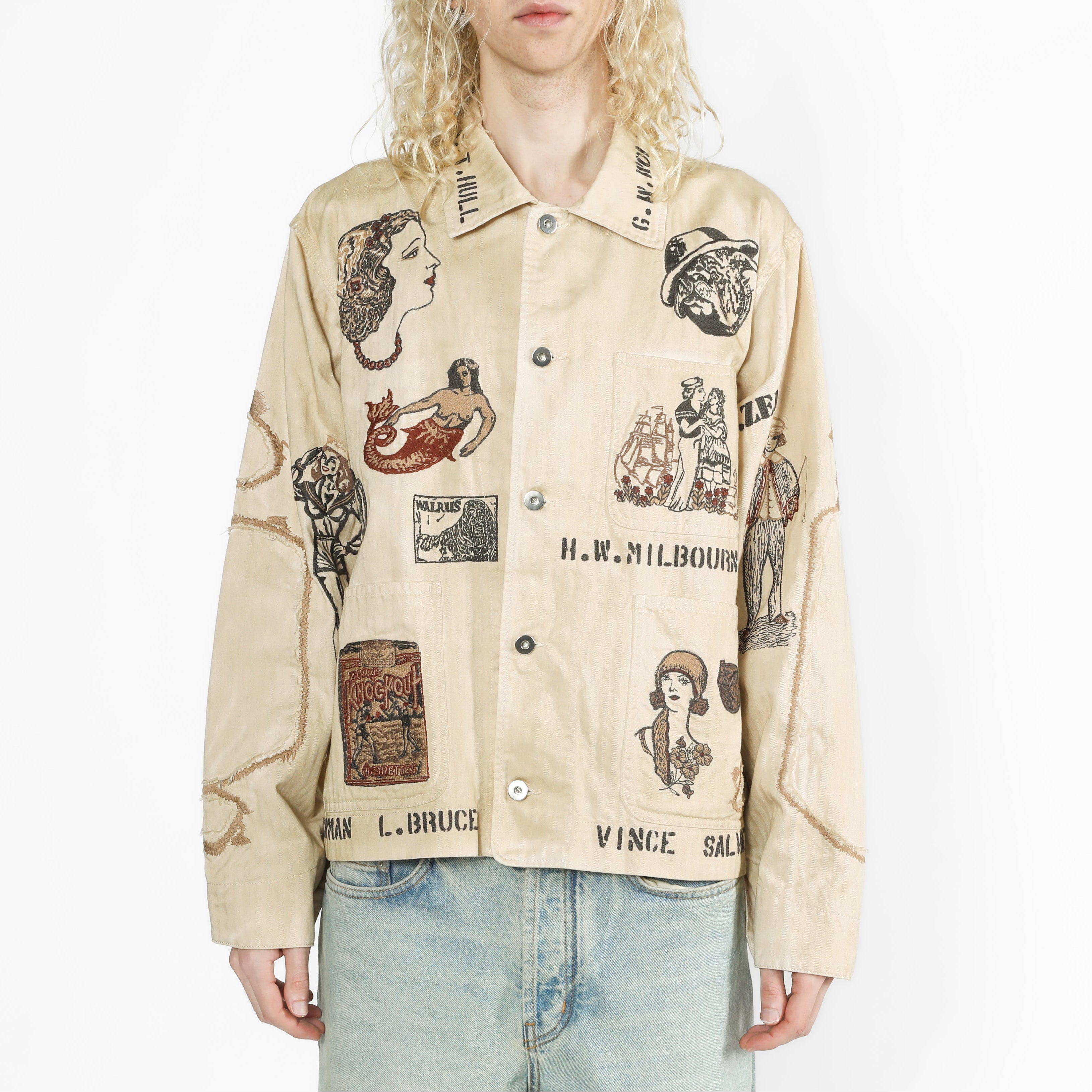 A model with long blonde hair wearing a beige button-up shirt jacket covered in vintage-style illustrated patches and text, paired with light blue jeans, against a plain white background.