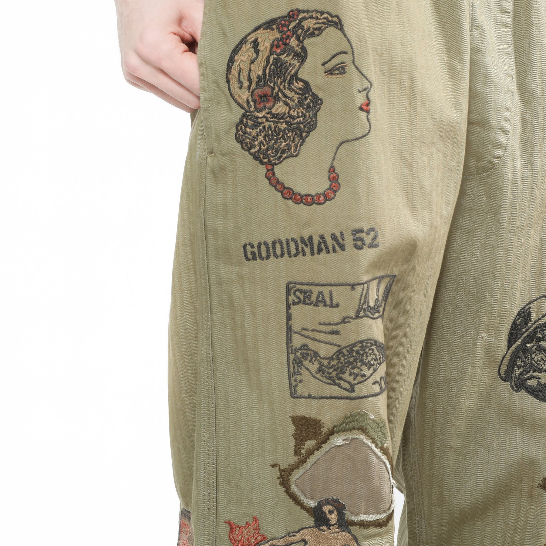 Close-up of a pair of olive green pants with vintage-style embroidered graphics and text including 'GOODMAN 52' and 'SEAL'.
