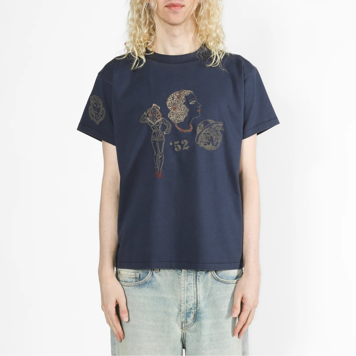 A model with long blonde hair wearing a navy blue graphic t-shirt and light wash jeans, standing against a plain white studio background.