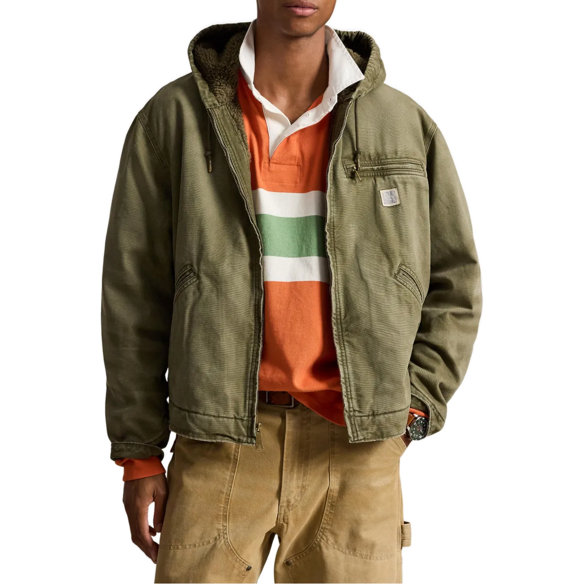 A man wearing an olive green hooded utility jacket over an orange and green striped polo shirt, paired with tan cargo pants, against a plain white background.