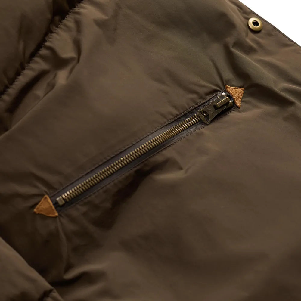 Close-up of a dark brown puffer jacket showing a diagonal zippered pocket with tan leather accents and brass snap buttons.