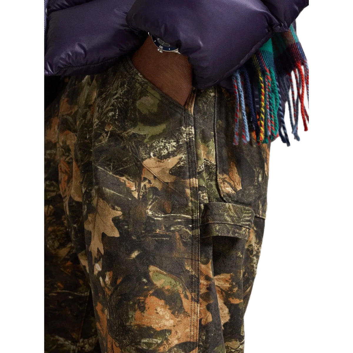 Close-up of a person wearing camouflage pants with a dark navy puffer jacket and a multicolored fringed scarf, against a white background.