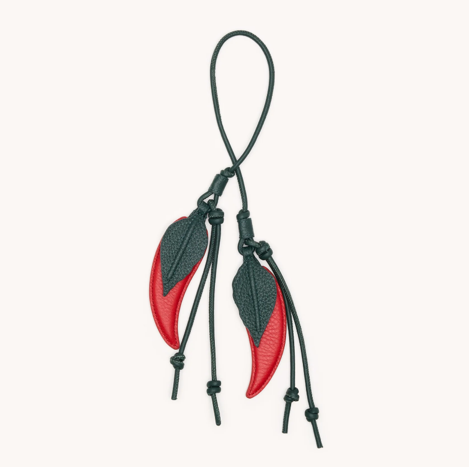 A pair of red and dark green leather chili pepper-shaped bag charms with dark green cords, shown on a plain off-white background.