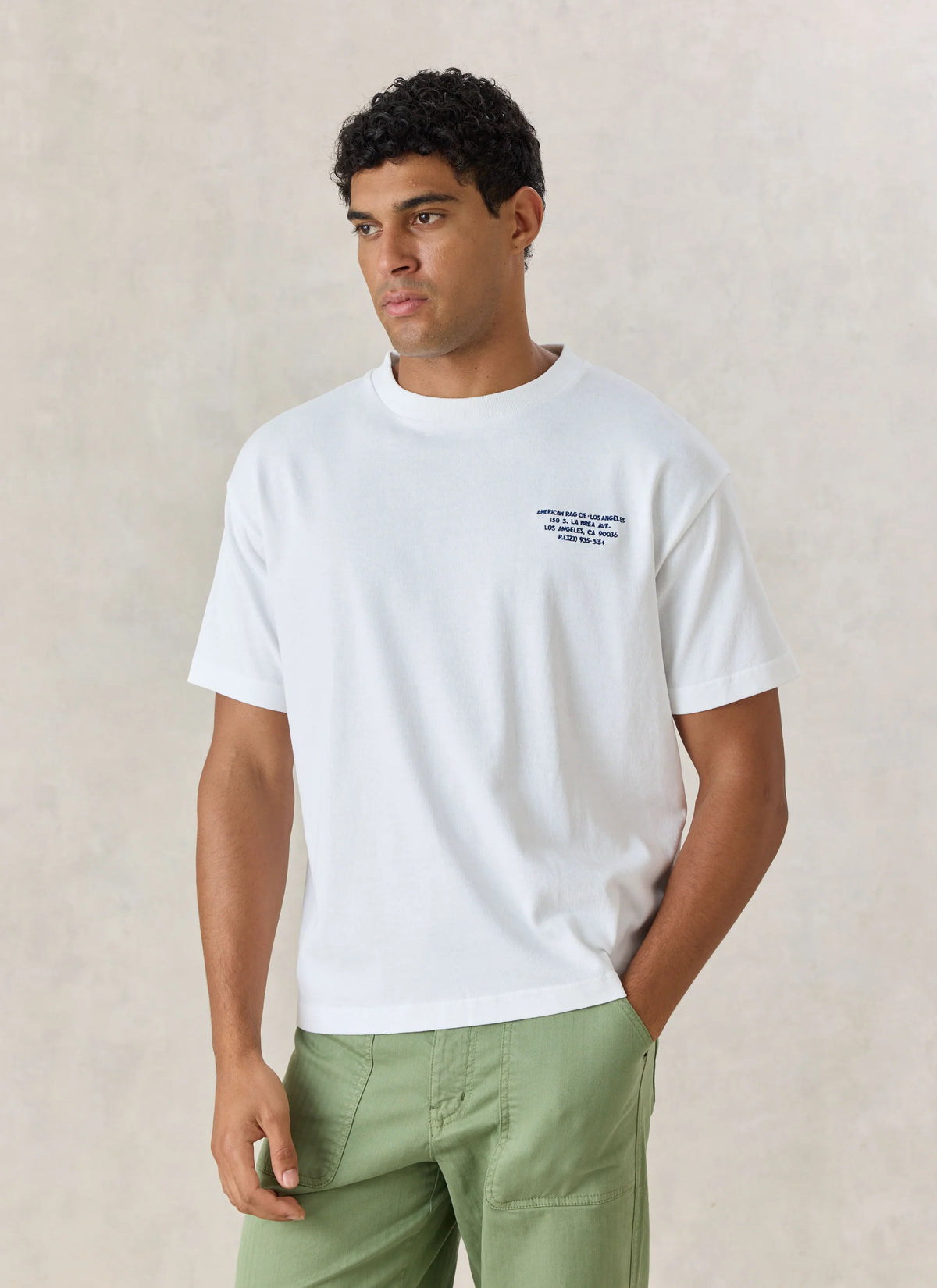 A man wearing a white short-sleeve crewneck t-shirt with a small navy blue graphic on the left chest, paired with sage green pants, standing against a textured beige background.