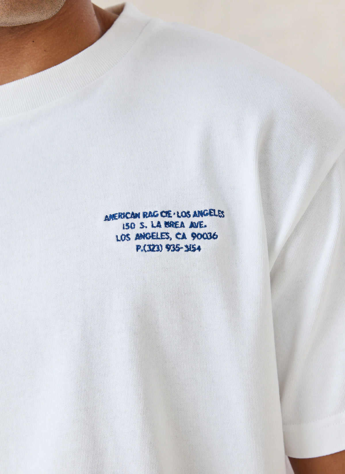 Close-up of a white short-sleeve crewneck t-shirt with blue embroidered text on the left chest reading 'AMERICAN RAG CIE•LOS ANGELES 150 S. LA BREA AVE.