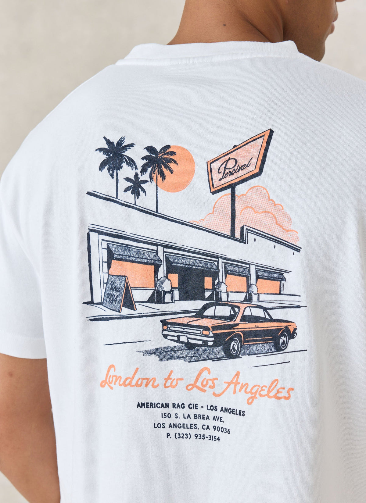 A person wearing a white graphic T-shirt, shown from the back. The shirt features a retro-style illustration of a building with palm trees, a sunset, a vintage car, and orange text reading 'London to Los Angeles' with smaller address text below.