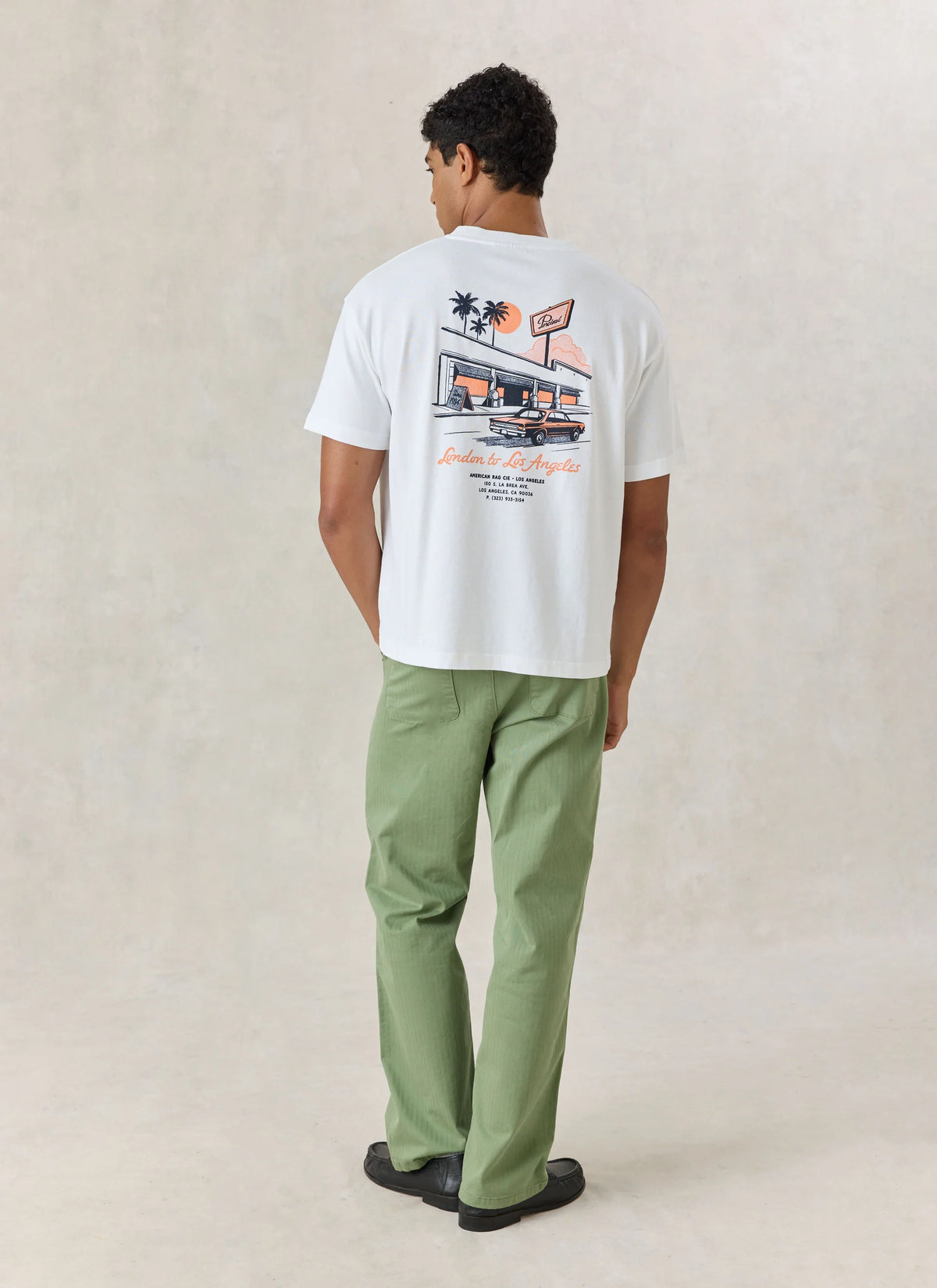 A man stands with his back to the camera wearing a white graphic t-shirt with a retro design on the back, paired with sage green pants and black shoes, against a textured beige studio backdrop.