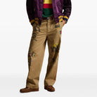 Person wearing a colorful jacket, striped shirt, and beige pants with patches on a white background