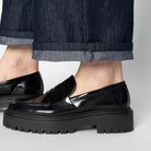 Black glossy loafers worn with rolled-up jeans on a light gray background
