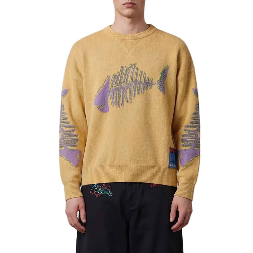Man wearing a mustard yellow sweater with abstract designs on sleeves and black pants against a plain background