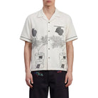 Man wearing a white patterned shirt and black pants standing against a plain background