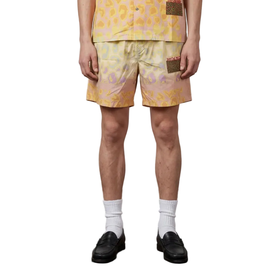 Man wearing a yellow patterned shirt and shorts standing against a plain background