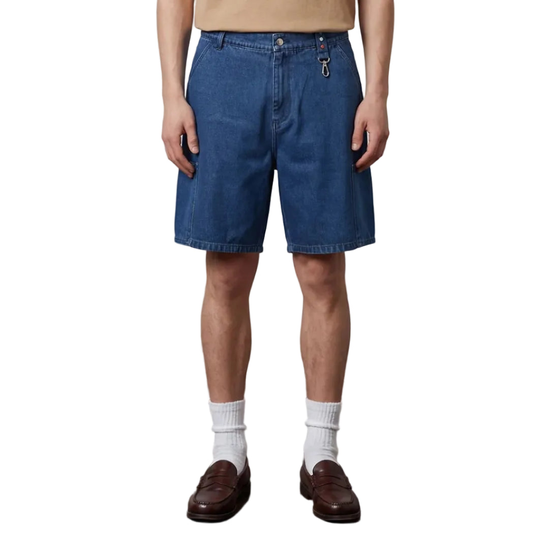 Man wearing a beige t-shirt and blue shorts standing on a wooden floor with a plain background