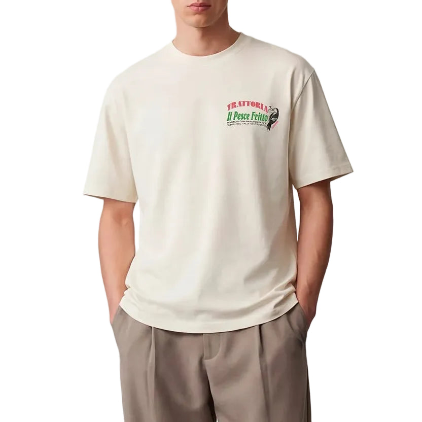 Man wearing a beige t-shirt with a logo and beige pants standing against a plain background