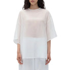 A model stands against a plain white studio background wearing a sheer white short-sleeve top with a wide round neckline and loose fit, paired with white wide-leg cropped pants.