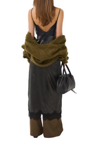 A woman is shown from behind wearing a black lace-trimmed maxi dress, a dark camisole, and holding an olive green fuzzy cardigan and a black handbag against a plain white background.