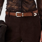 A close-up of a person wearing a dark brown lace and suede top with matching dark brown trousers, styled with a brown crocodile-embossed belt and a silver buckle.
