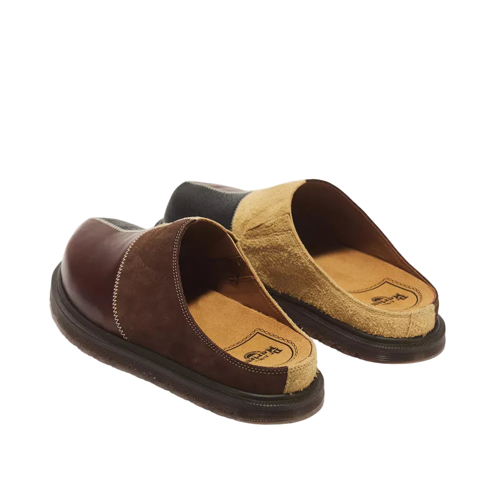 A pair of slip-on mule slippers on a white background. The slippers feature a dark brown leather upper, a tan suede-like side panel, and a black sole.
