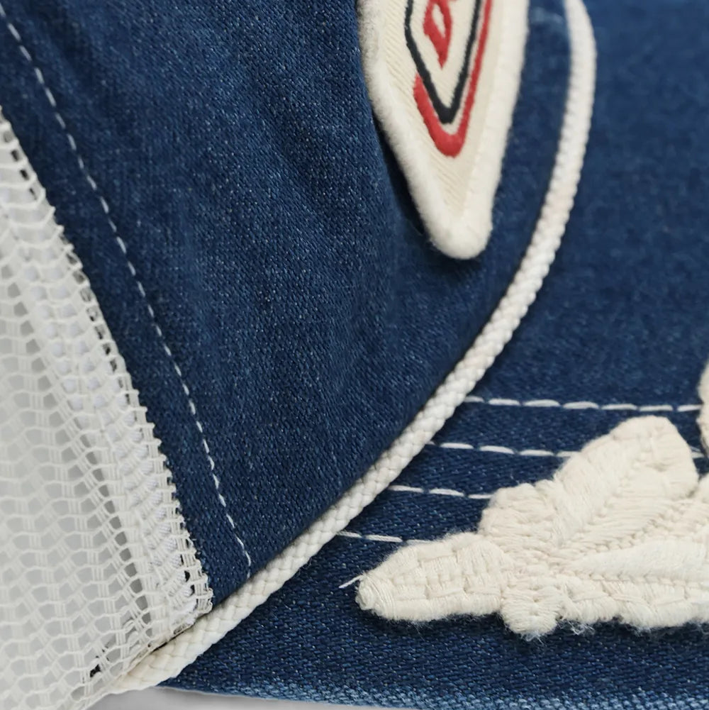 Close-up of a blue denim trucker cap with white mesh side panels, white contrast stitching, and a white embroidered patch featuring red and black lettering.