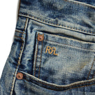Close-up of a blue denim jeans pocket showing the embroidered 'RRL' logo in gold on the back pocket, with copper-toned rivets and a white background visible on the left.