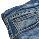 Close-up of a blue denim jeans pocket showing the back of the waistband, metal rivets, and a gold embroidered 'RRL' logo on the pocket.