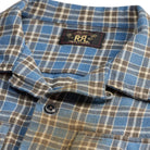 Close-up of a plaid button-up shirt with a blue, brown, and beige pattern, featuring a collar label with 'R.A.C' branding.