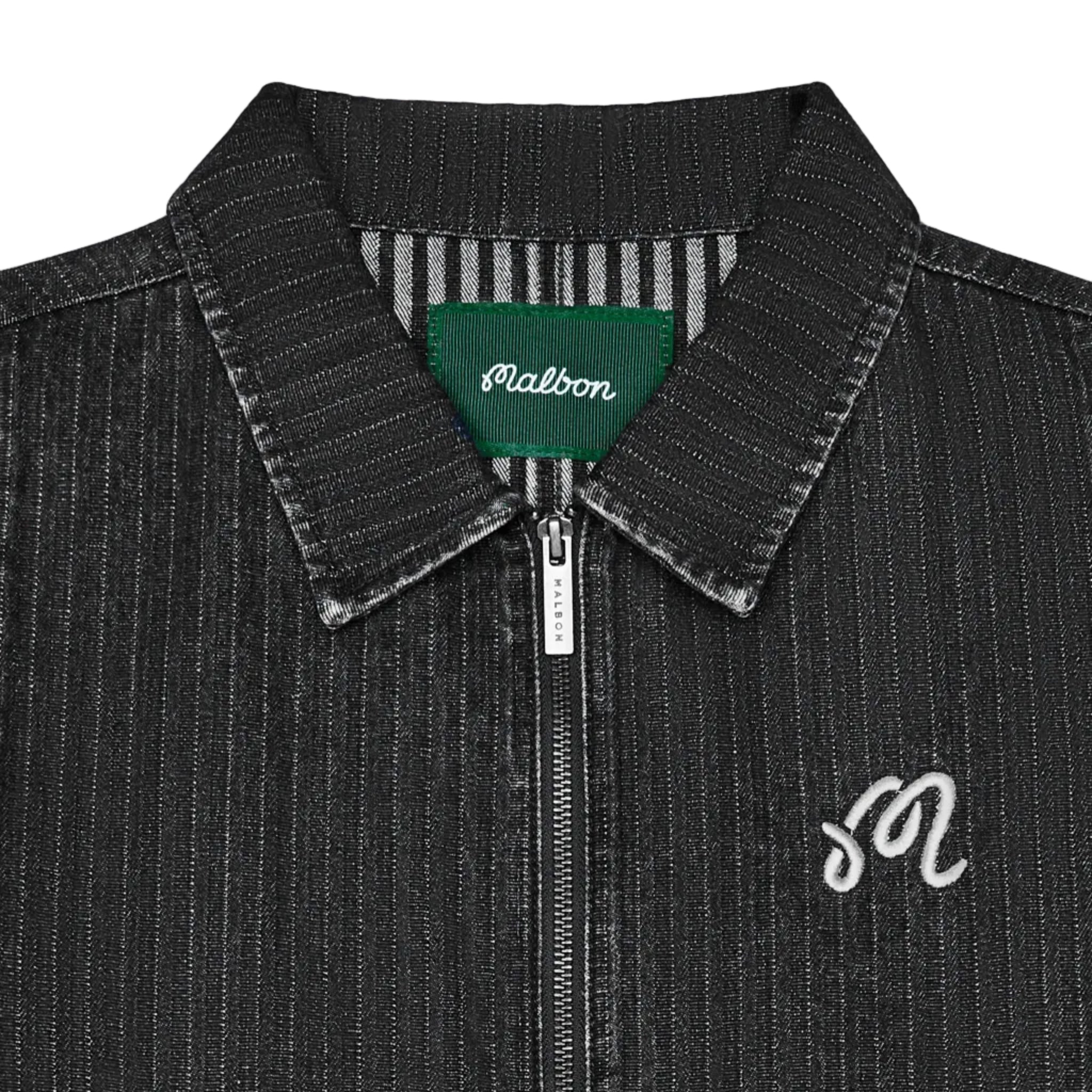 Close-up of a dark gray textured zip-up jacket with a pointed collar, featuring a green Malbon neck label and a white embroidered script 'M' on the chest.