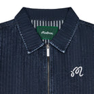 Close-up of a dark blue denim zip-up jacket with a pointed collar, featuring a green Malbon neck label and a white embroidered script 'M' on the chest.