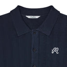 Close-up of a dark navy blue textured knit polo shirt with a white embroidered script logo on the chest and a visible 'Malbon' brand label inside the collar.