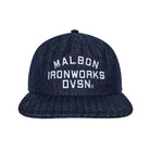 A dark navy blue denim flat-brim cap with white embroidered text reading 'MALBON IRONWORKS DVSN.' on the front, shown against a plain white background.