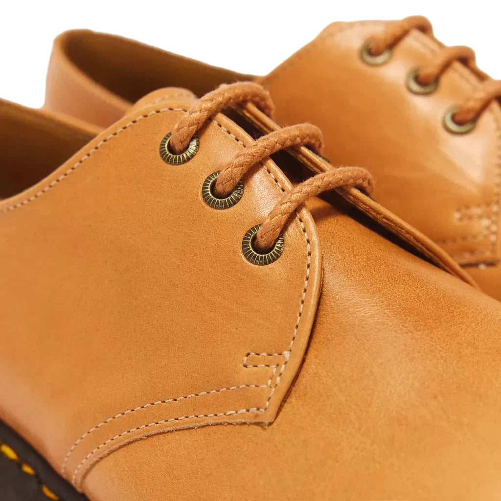 A close-up shot of a tan leather lace-up shoe, showing the textured leather upper, decorative stitching, brass eyelets, and a black sole with yellow stitching.