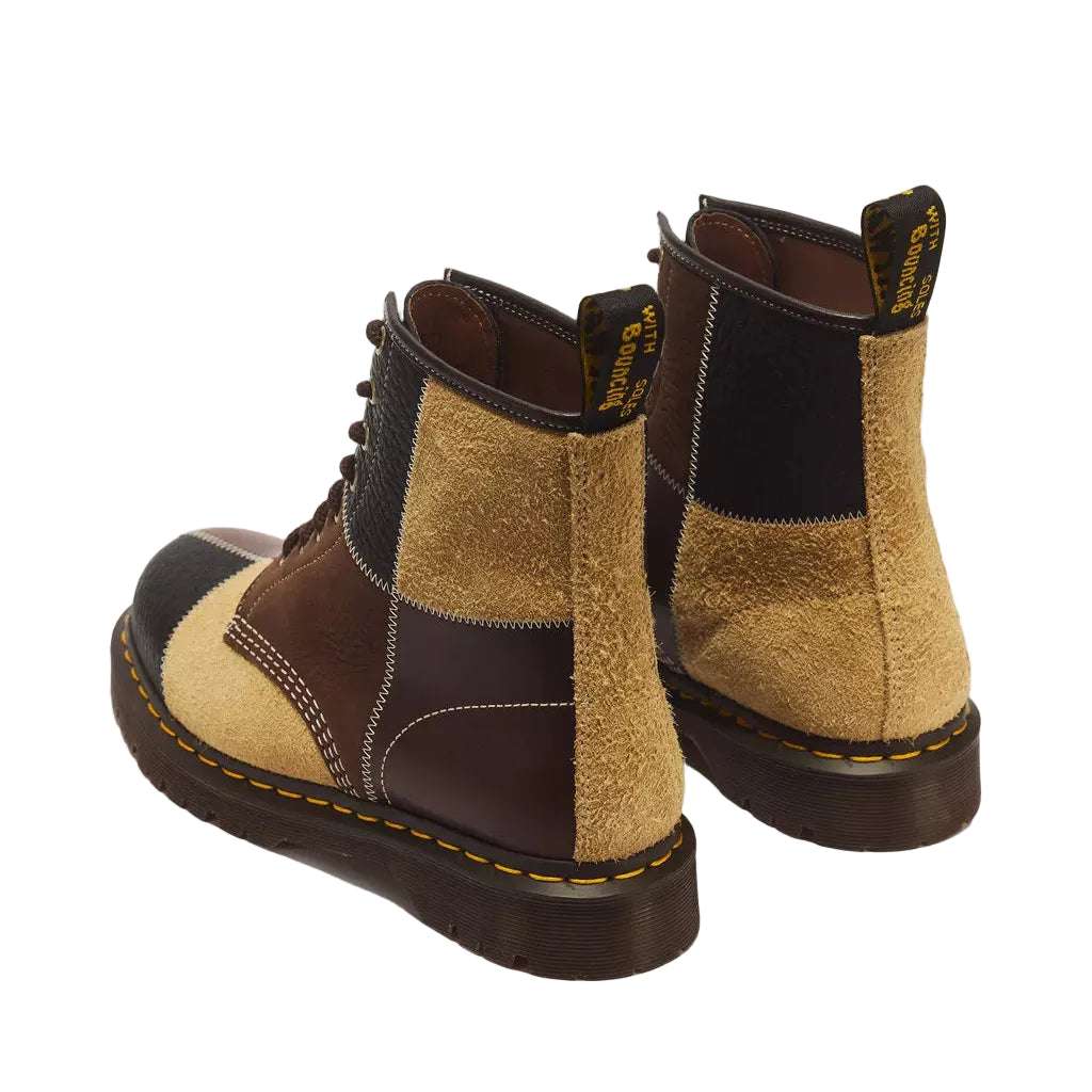 A pair of Dr. Martens ankle boots shown from a rear three-quarter angle, featuring a colorblock design with tan suede, dark brown leather, and black panels, finished with a dark brown sole and yellow stitching.