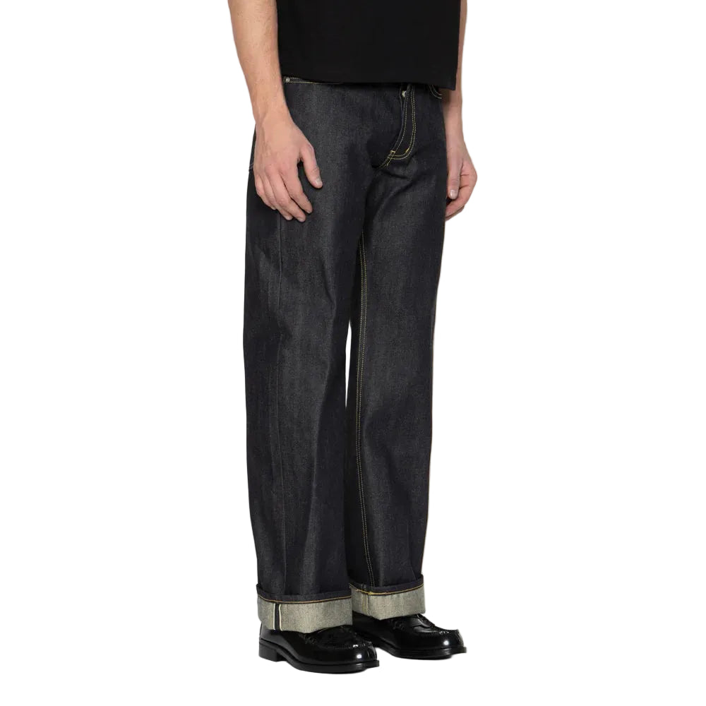 A person wearing dark indigo denim jeans with a rolled cuff showing a lighter inner lining, paired with a black shirt and black leather shoes, against a white background.