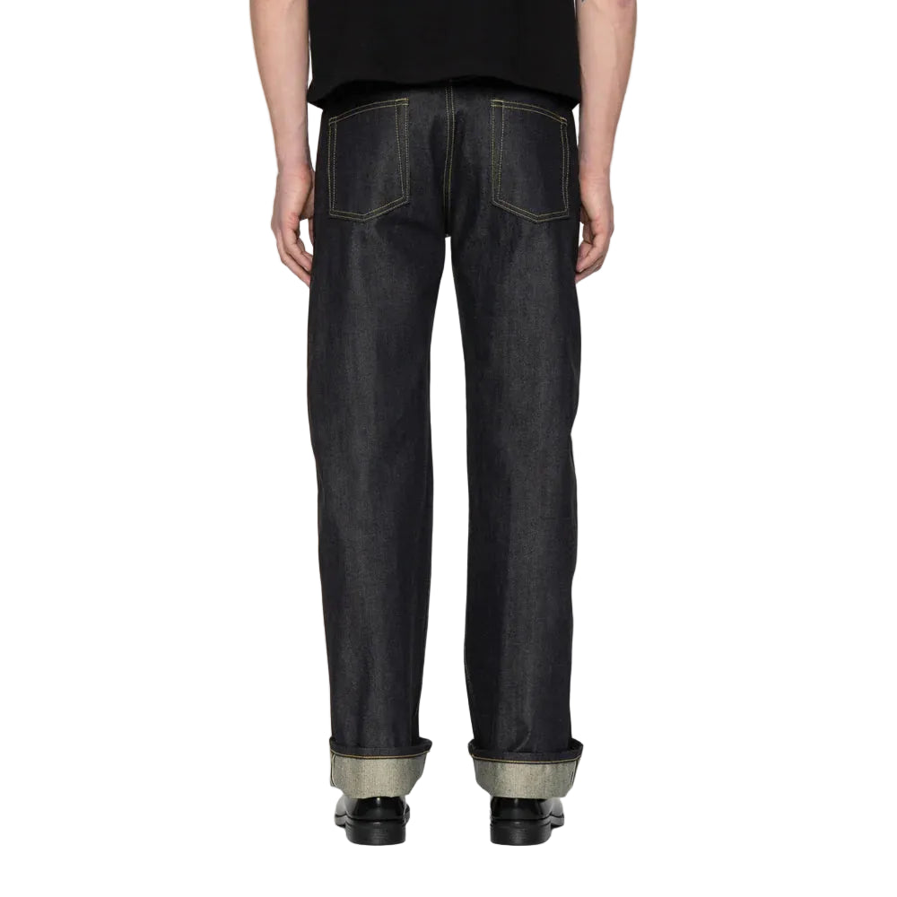 Rear view of a person wearing dark indigo denim jeans with a black shirt and black shoes, standing against a white background.