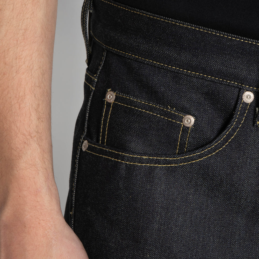 Close-up of a person wearing dark indigo denim jeans, showing the back pocket with contrast yellow stitching and silver metal rivets.
