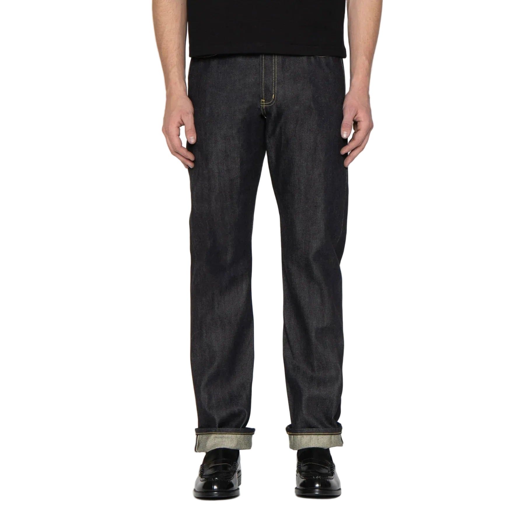 A person wearing dark indigo straight-leg jeans with rolled cuffs, paired with a black shirt and black shoes, standing against a white background.