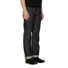 A person wearing dark indigo denim jeans with rolled cuffs showing a light inner lining, paired with black leather shoes, photographed against a white background.