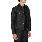 A man wearing a dark indigo denim jacket over a black shirt and matching dark jeans, standing against a plain white background.