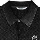 Close-up of a black ribbed knit polo shirt featuring a white embroidered monogram logo on the chest and a visible 'Malbon' brand label inside the collar.